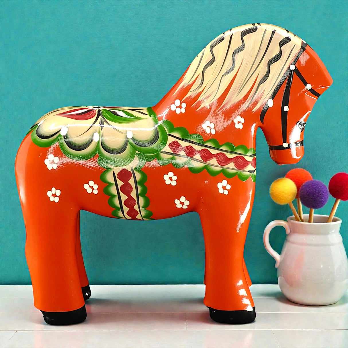 A handcrafted red Swedish Dala Horse "Färnäshästen" by Nils Olsson (14 cm) stands beside a white vase filled with colorful felt balls on sticks, set against a teal background.