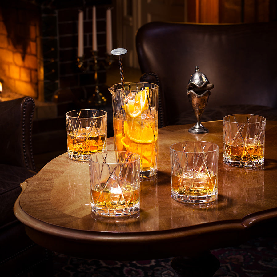 A wooden table features five glasses of amber whiskey, one with a stirrer and citrus slices, accompanied by a silver container. Orrefors City Old Fashioned Glass 8oz (2-Pack) gleams warmly against the fireplace in the background.