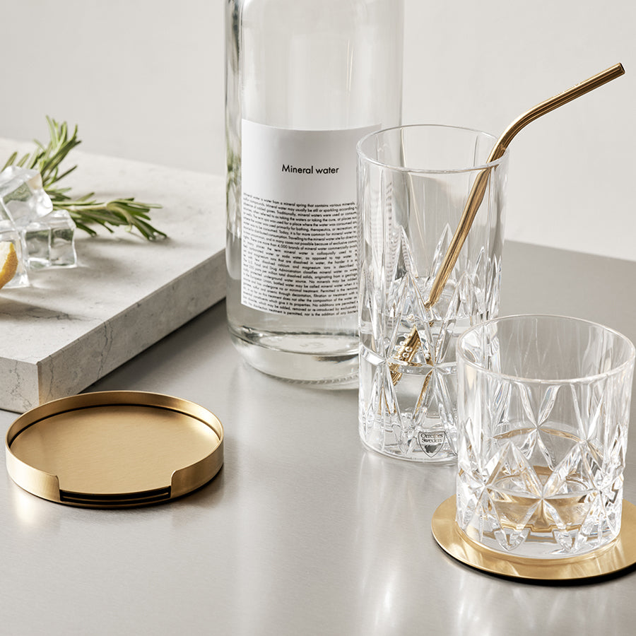A bottle of mineral water, two Orrefors: Peak Old Fashioned Glasses from the 4-Pack by Martti Rytkonen—one with a gold straw—plus gold coasters on a gray surface, with a tray of ice and herbs in the background.