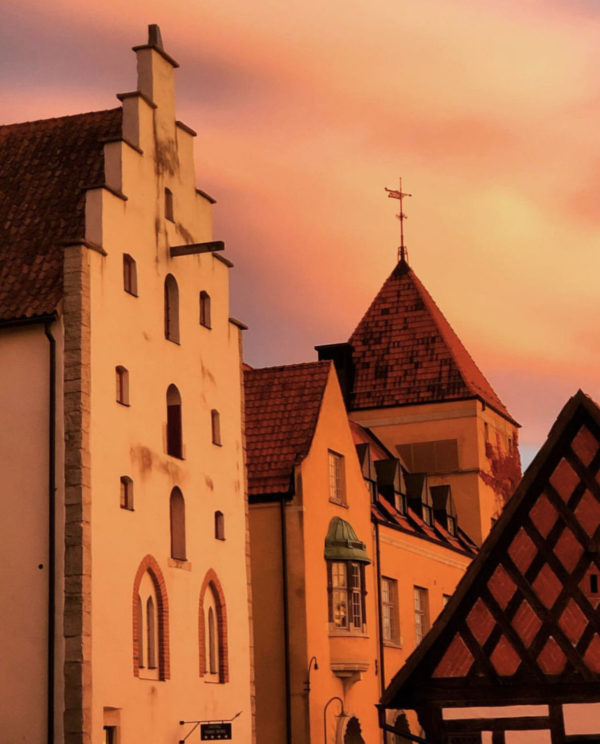 The Visby blanket in rust-colored wool by Kerstin Landström Design captures the essence of Swedish heritage, reminiscent of historic European buildings with steep roofs set against a warm, orange sunset sky, blending seamlessly with the scene's timeless charm.