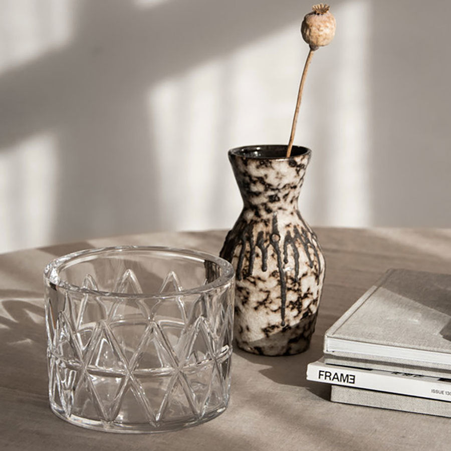 An Orrefors: Peak Bowl with a textured design, a small ceramic vase holding a dried flower, and two stacked books are placed on a table under soft lighting.