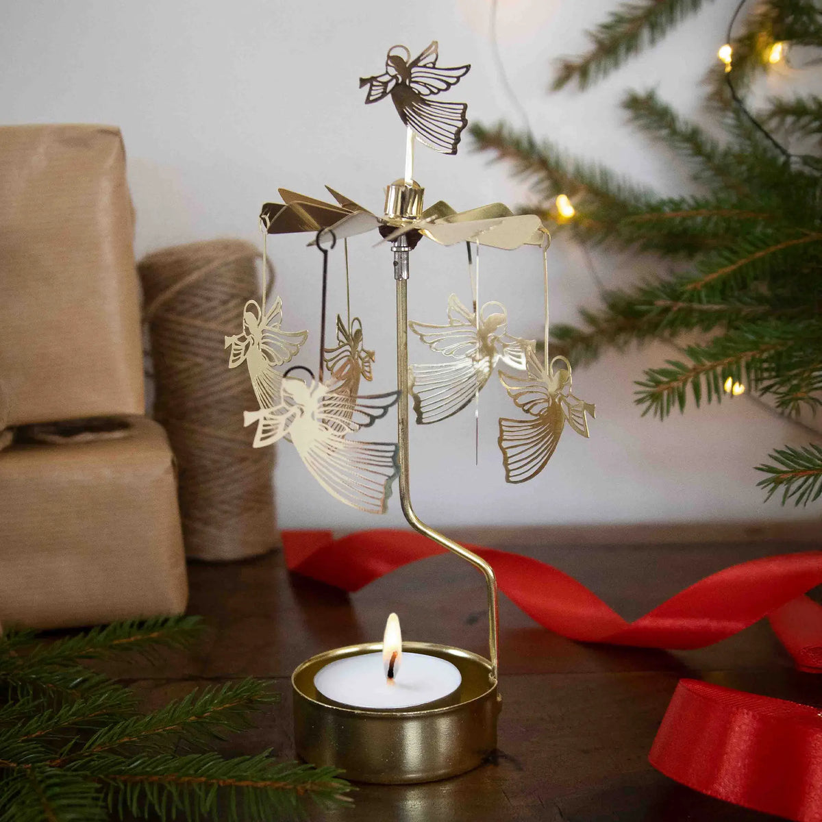 Gold angel rotary candle holder spinning above lit tealight, with pine branches, wrapped gifts, twine, and red ribbon arranged on a decorated table.