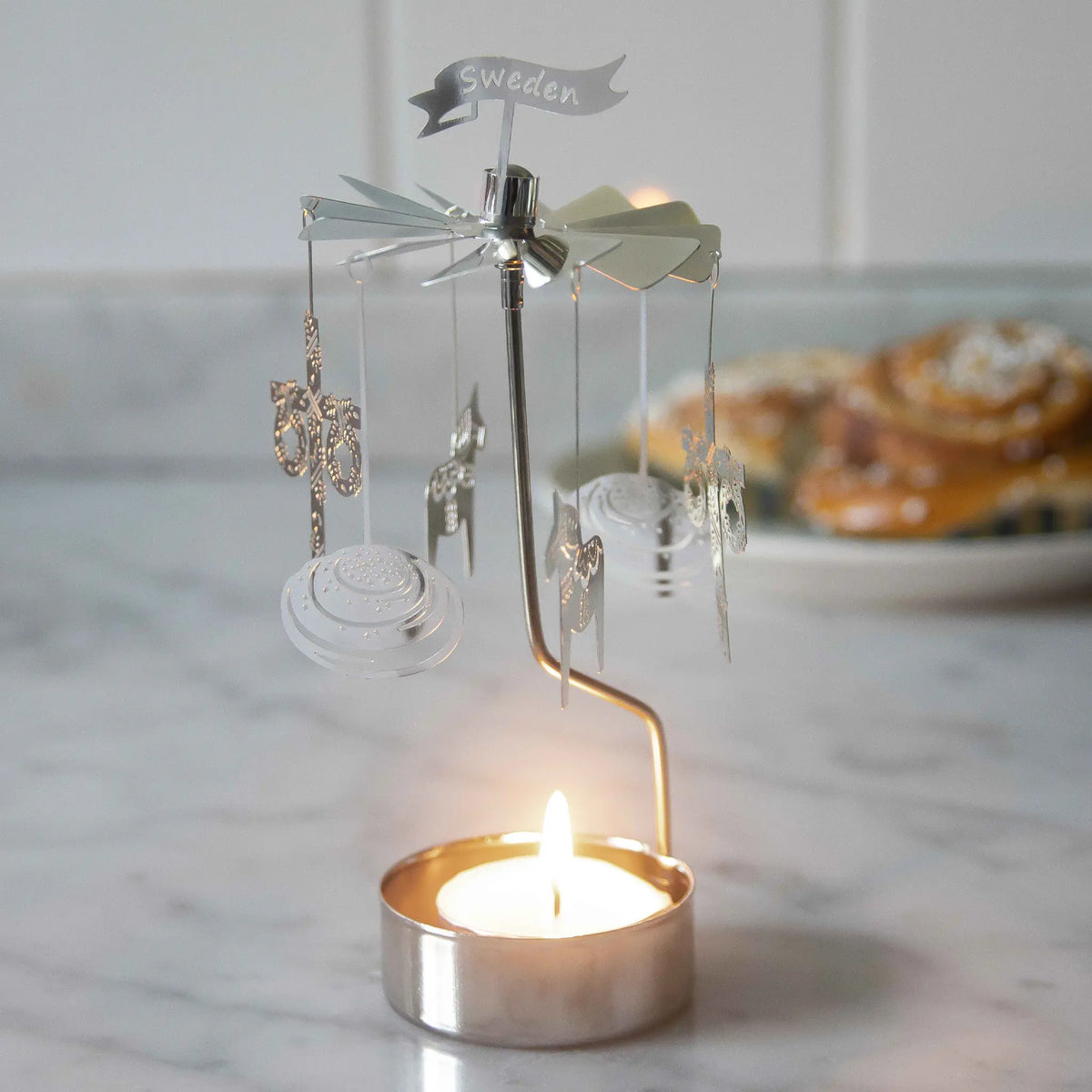 Rotary candle holder with hanging ornaments spinning above a lit tealight on a marble counter, cinnamon buns blurred in the background.