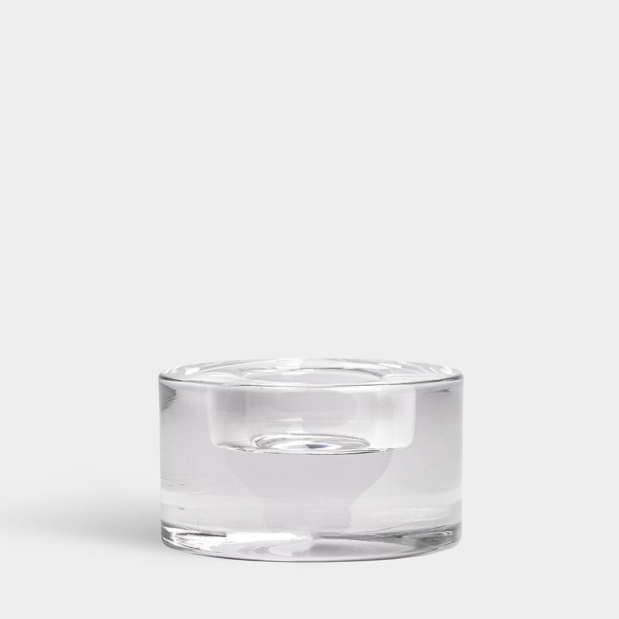 The Orrefors: Puck Votive Medium Candle Holder, a stunning clear glass piece designed by Lena Bergström, stands elegantly against a plain, light gray background.