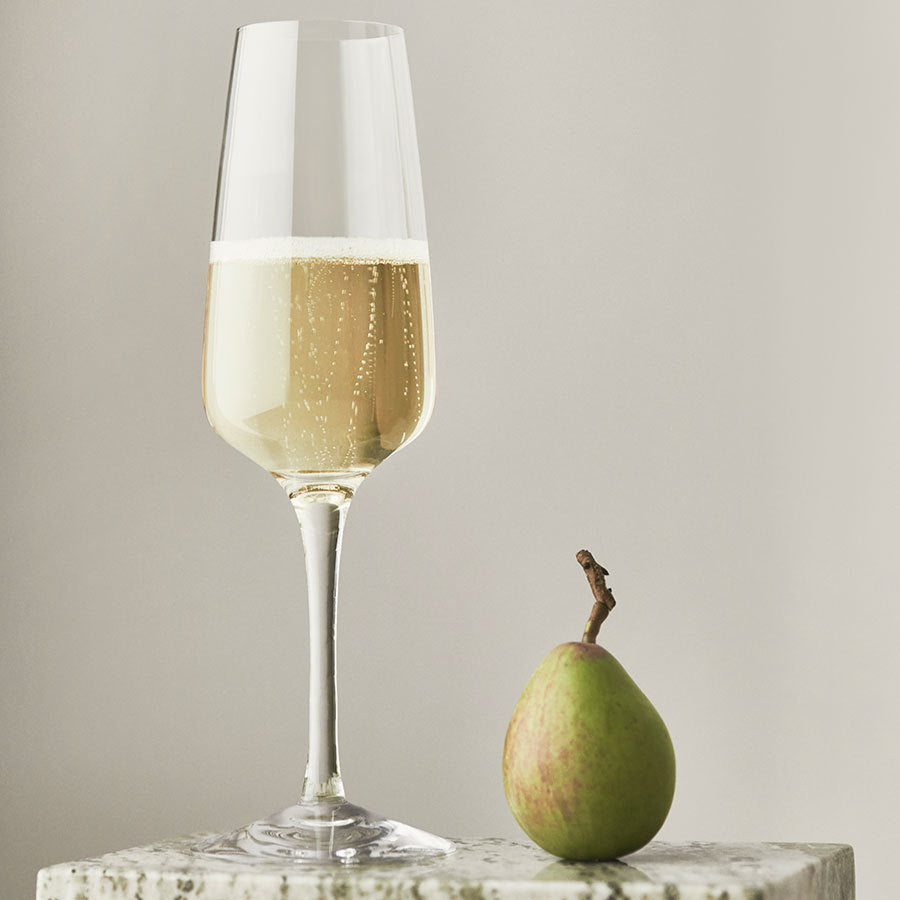 An Orrefors: Pulse Champagne Glass holds sparkling wine, perfectly paired with a lone pear on sleek marble.
