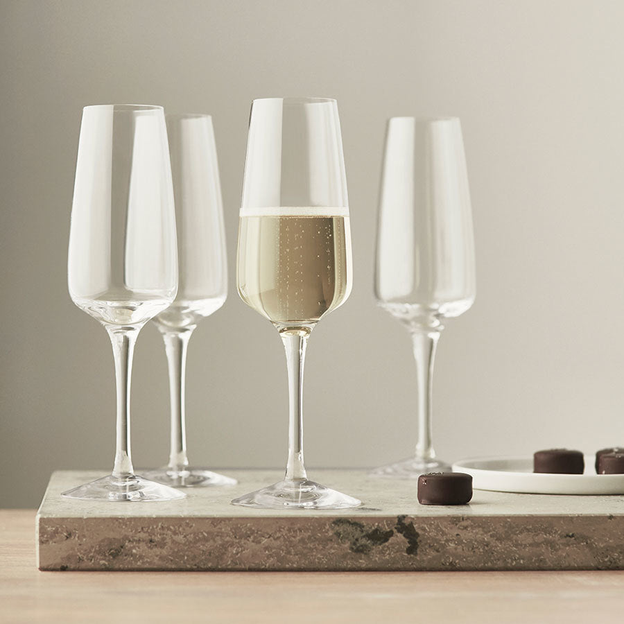 Four Orrefors Pulse Champagne Glasses sit on a marble tray, one filled with bubbly. A nearby plate of chocolates enhances this image of indulgence and showcases the refined elegance of the Pulse Collection.