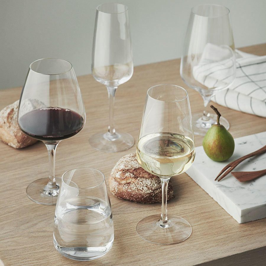 A wooden table showcases the Orrefors Pulse Collection with five distinct wine glasses and a sleek champagne glass. Red or white wine fills some glasses, while others are empty. A glass of water, a pear, bread rolls, and a folded cloth napkin complete the display.