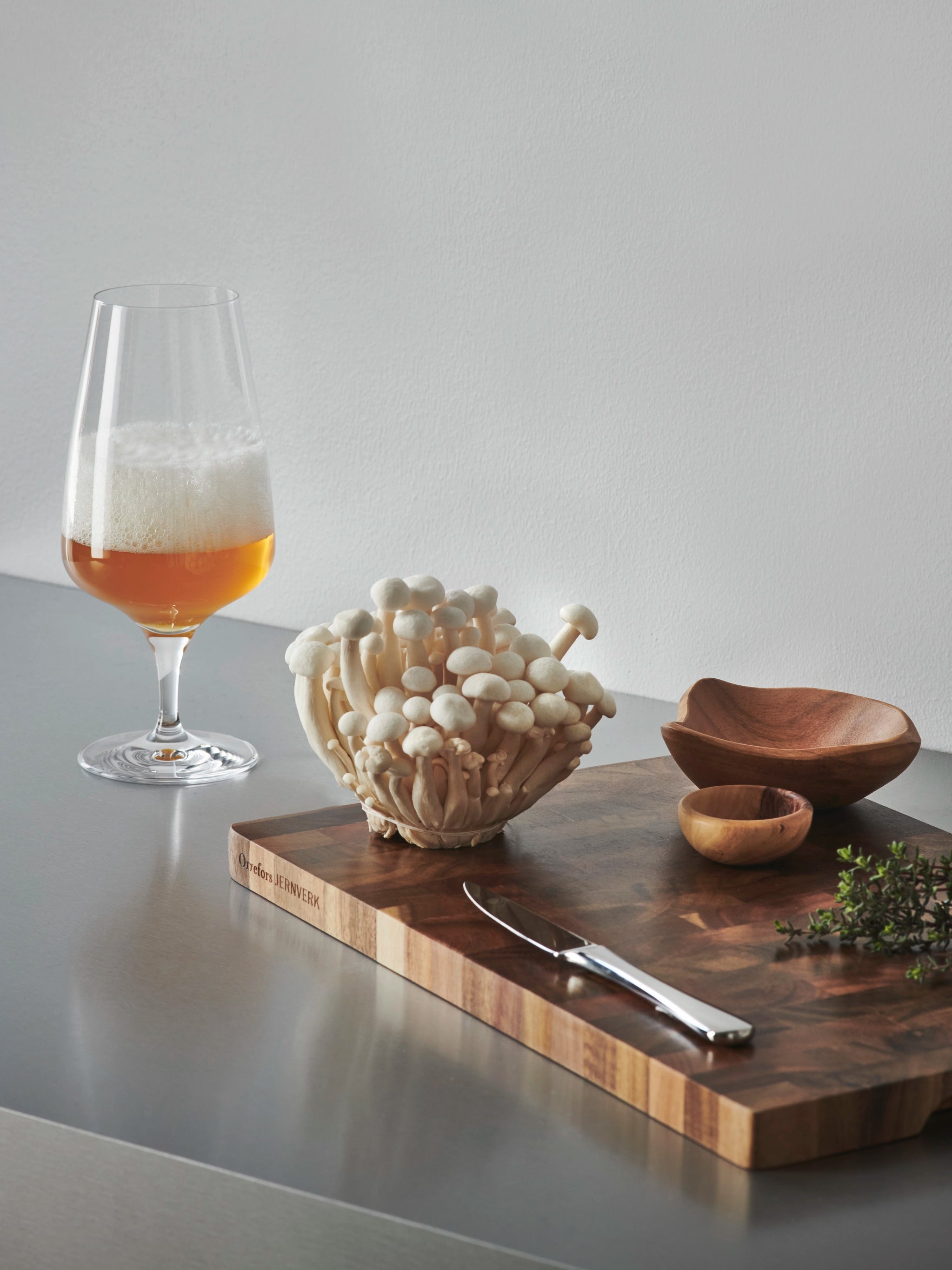 A glass of beer in an Orrefors: Pulse Beer Glass 18.5oz (4-Pack) by Ingegerd Raman sits alongside white mushrooms, a knife, wooden bowls, and fresh herbs, showcasing Scandinavian simplicity on the counter.