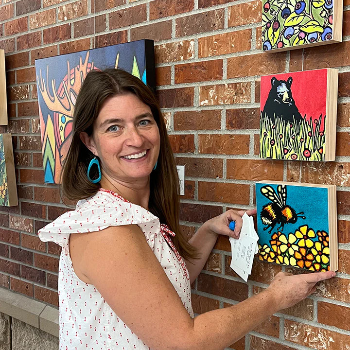 A woman smiles while hanging the vibrant "Nature's Notes 1000 Piece 'Mixed Up!'" PuzzleTwist jigsaw puzzle by Sarah Angst on a brick wall among other colorful animal artworks, featuring the distinctive Maynards style.
