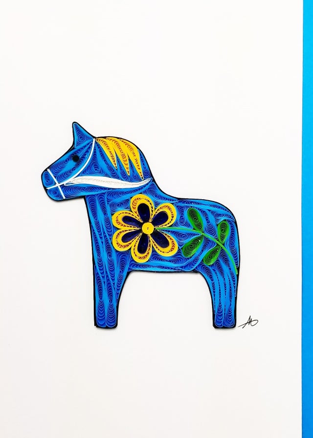 The "Cards: Dala Horse Blue Quilling" features a paper-quilled Dala horse adorned with a striking yellow and black Swedish flower, accented by rich green leaves on its side.