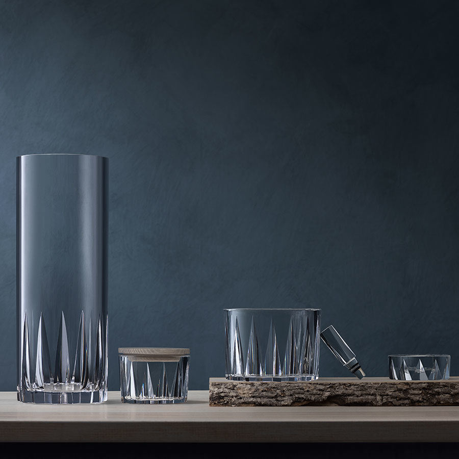 A set of clear glassware, inspired by Swedish nature, rests on a wooden surface against a dark backdrop. It includes a tall vase, a lidded jar, a low vessel on a rustic tray, and an elegant Orrefors: Sarek Bowl with Lid.