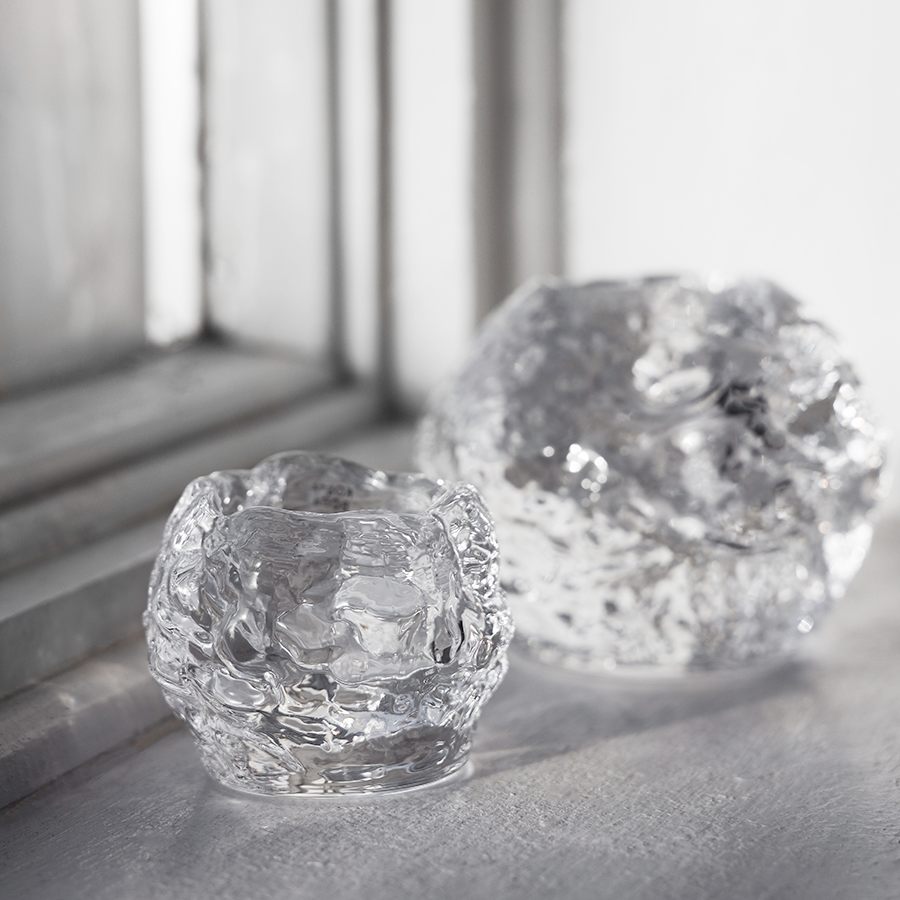 Two clear, textured KostaBoda: Snowball Votive Small 60mm candle holders by Ann Warff sit on a white surface near a window, light reflecting off their uneven surfaces.