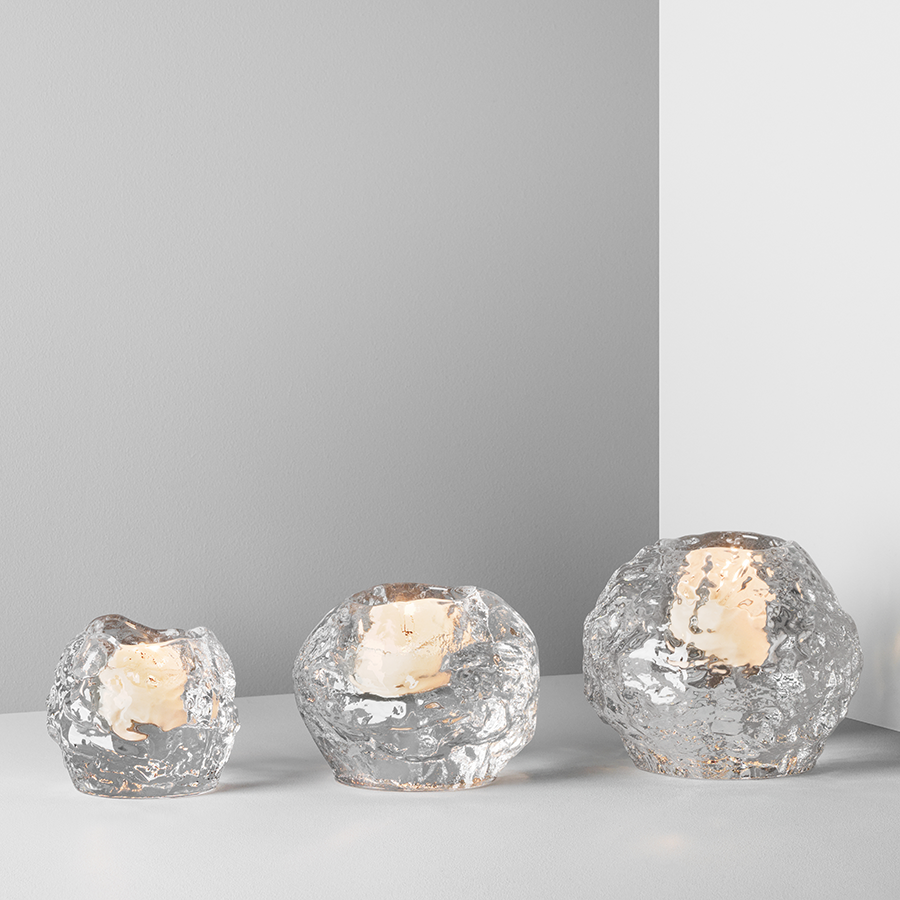 Three glass candle holders, including the KostaBoda Snowball Votive Small 60mm by Ann Warff holding lit candles, are displayed on a light grey surface against a grey and white backdrop.