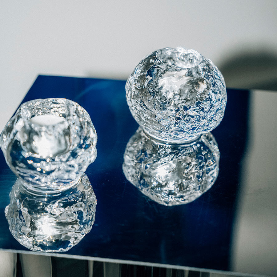 Two KostaBoda Snowball Votive Medium 70mm candle holders in clear textured glass sit on a reflective metallic surface, casting shimmering reflections and highlighting their intricate details.