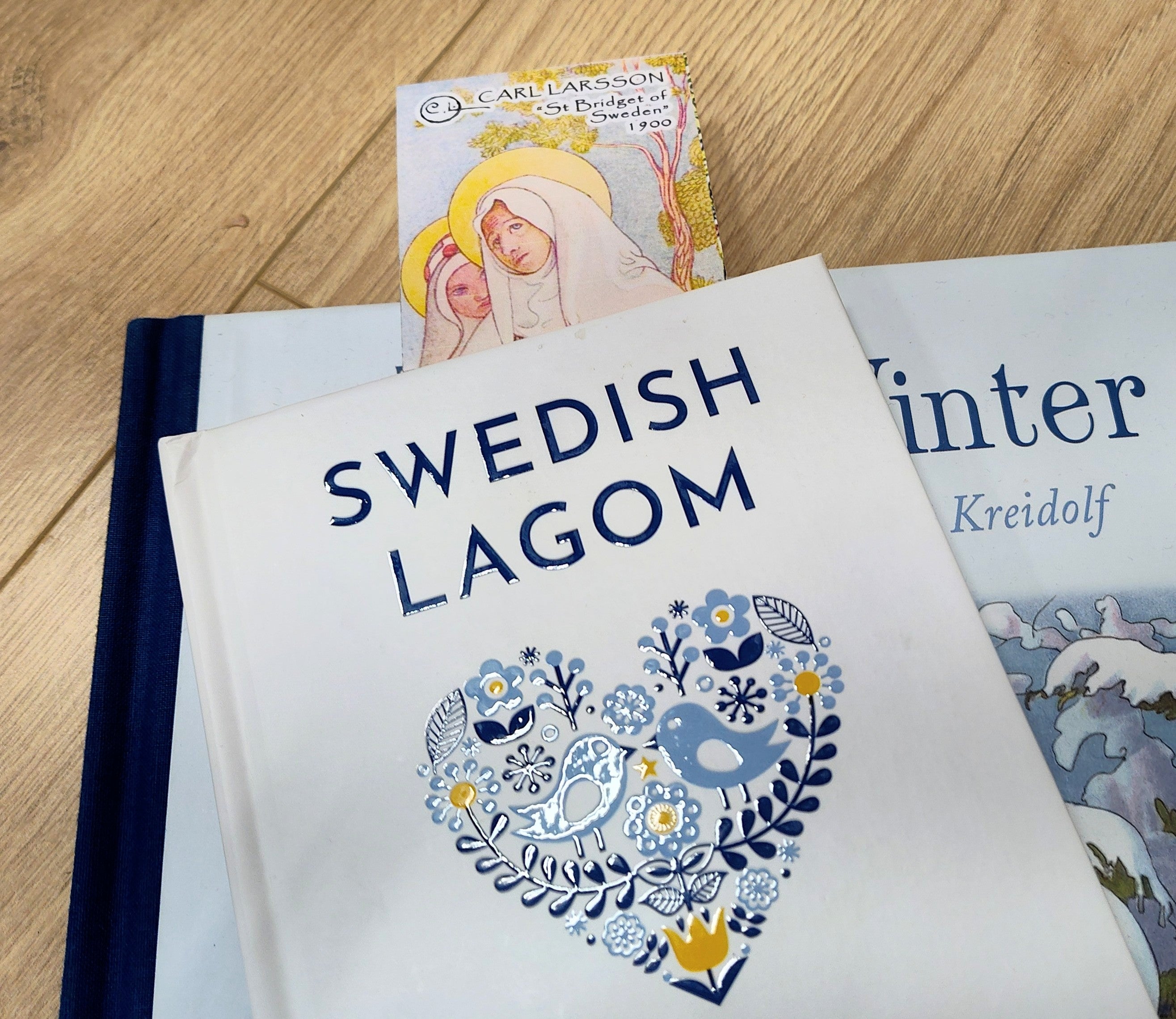 A book titled Swedish Lagom with a blue heart on the cover rests atop another, featuring a Carl Larsson “St. Bridget of Sweden” bookmark (8.5" × 2.5") that honors Scandinavian art.