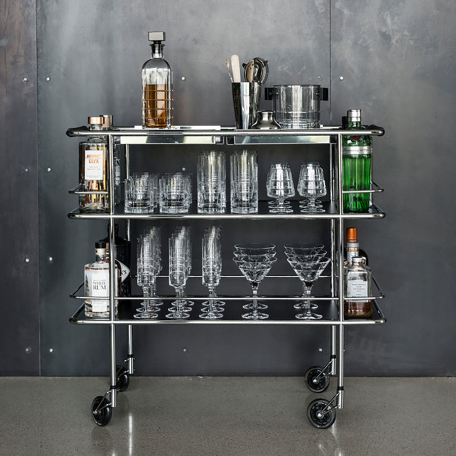 A metal bar cart with wheels displays various liquor bottles, Orrefors: Street Martini Glasses, and a cocktail shaker against a metallic wall backdrop.