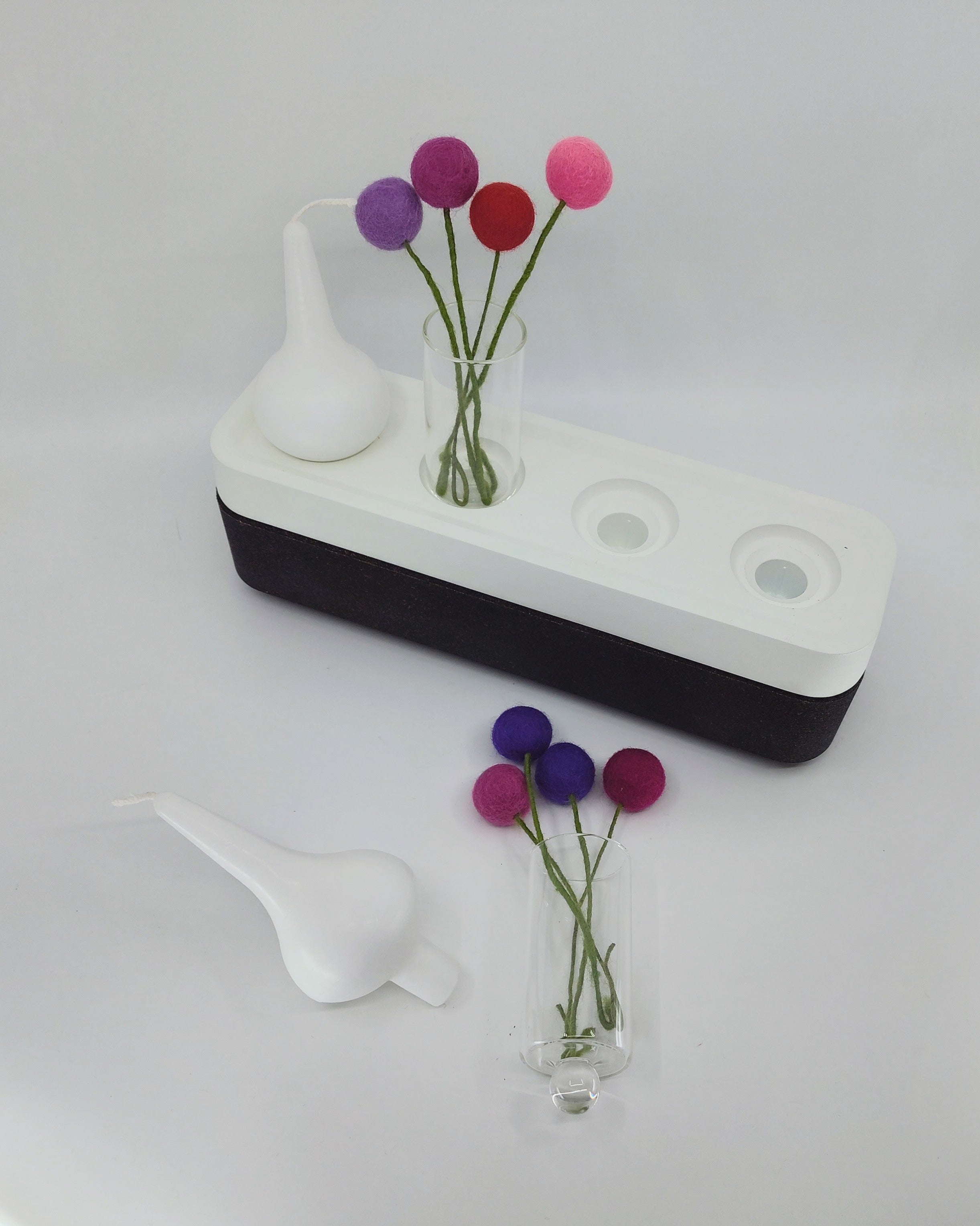 The Stumpastaken Swedish Candle Holder features a white container with a black base, a clear vase of colorful felt ball flowers, and a white funnel-like accessory, all set against a pristine white background to underscore its eco-friendly design.