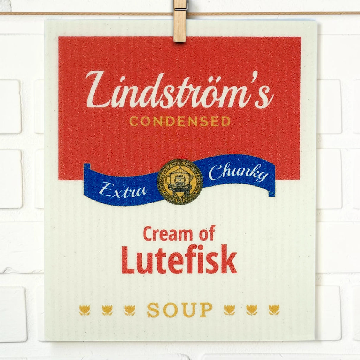 Swedish dishcloth designed like a soup can label with text: Lindströms Condensed Extra Chunky Cream of Lutefisk Soup, shown against a white brick background.