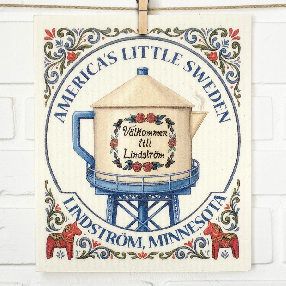 Decorative sign featuring the text “Americas Little Sweden, Lindström, Minnesota” with a teapot water tower, “Välkommen till Lindström,” and Swedish dishcloth floral patterns.
