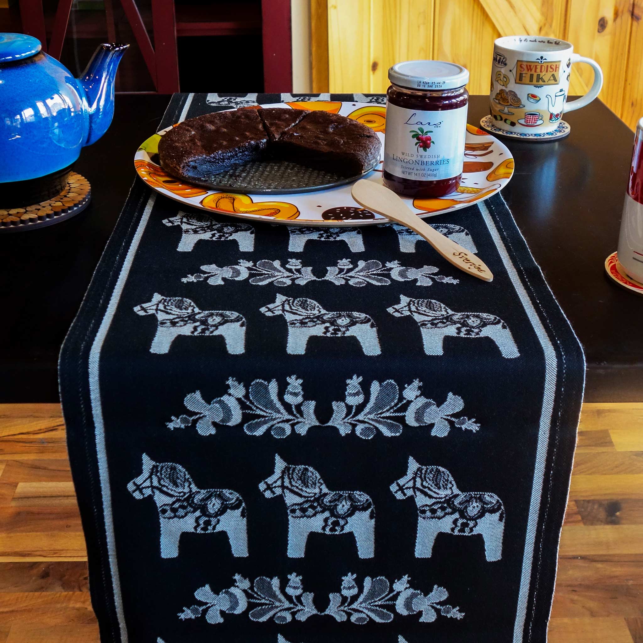 A table features a Siljan Grey runner with Swedish Dala Horses on black by Ekelund, adorned with a sliced chocolate cake on a decorative plate, a jar of lingonberry jam, and accompanied by a blue teapot and two cups.