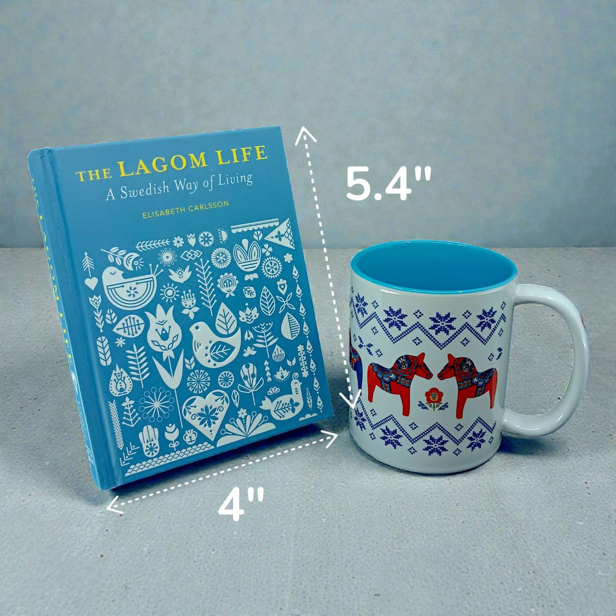 A blue book, "The Lagom Life: A Swedish Way of Living by Elisabeth Carlsson," measuring 4" x 5.4", stands beside a white mug featuring red horse designs.