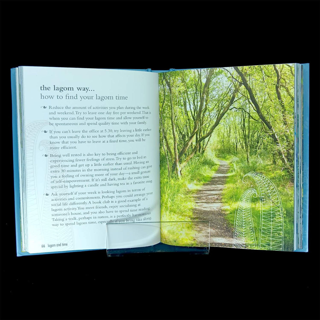 An open copy of "The Lagom Life: A Swedish Way of Living" by Elisabeth Carlsson displays tips on finding lagom time on the left page and a sunlit forest path lined with green trees on the right.