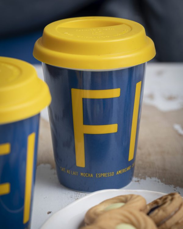 The Mug: "Fika" To-Go features a blue body, yellow lid, and bold yellow "FI" letters in takeaway style—an ideal Swedish souvenir for coffee lovers, shown beside cookies.