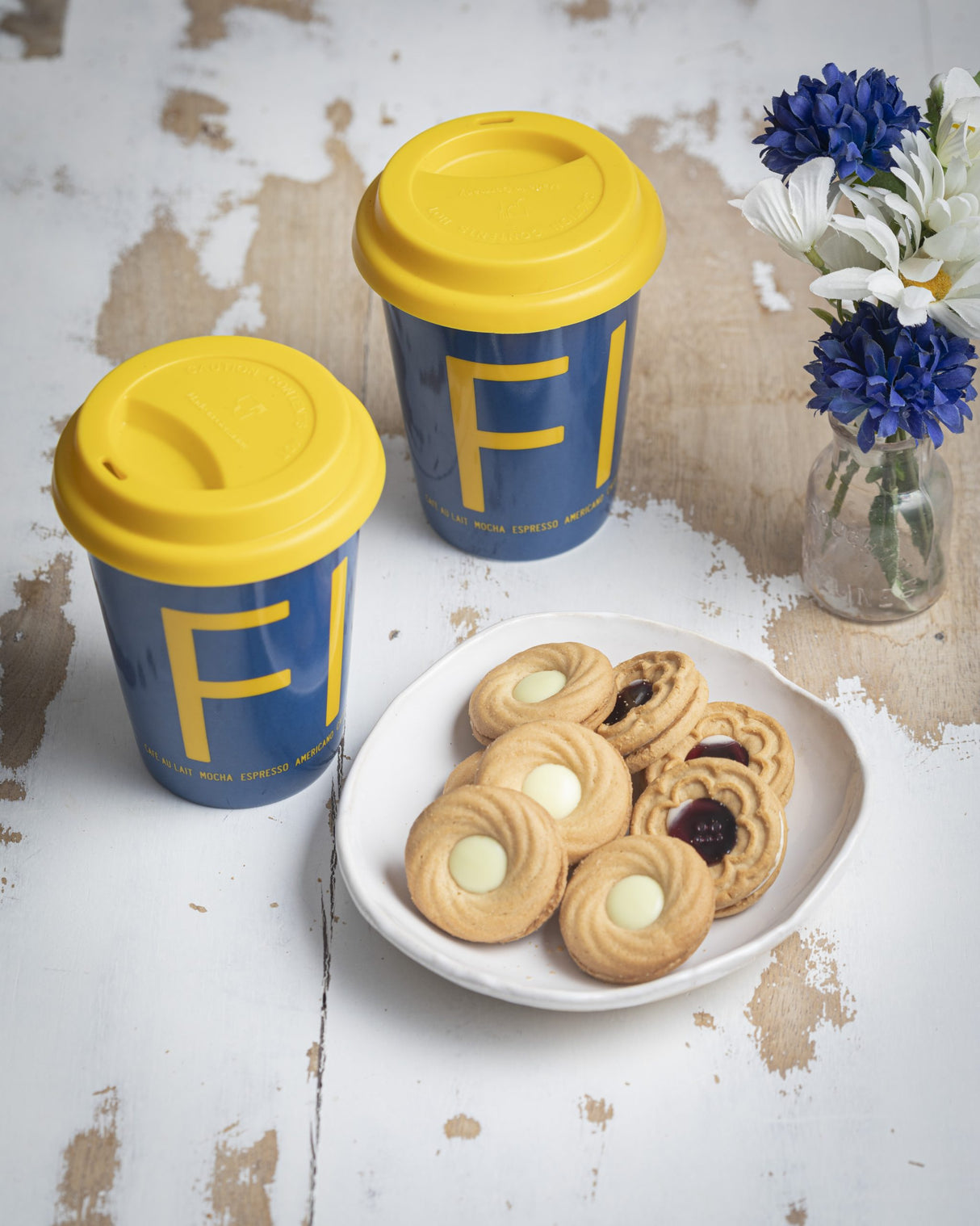 A "Fika" To-Go coffee mug with a yellow lid and blue sleeve sits next to a plate of assorted cookies and a small vase of blue and white flowers on a rustic white table.