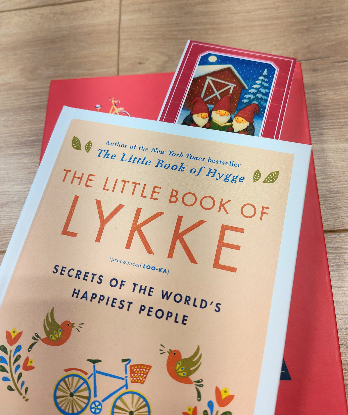 Two books rest on a wooden surface: “The Little Book of Lykke” atop a red, folklore-inspired volume with a barn and birds. A “Tomte Rules” bookmark (2.5" × 8.5") peeks out, lending Nordic winter charm to the scene.