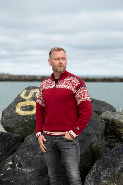 A person in a luxurious Arctic Circle Tor Merino wool sweater, colored a rich red, stands on rocks near a body of water, with a cloudy sky overhead.