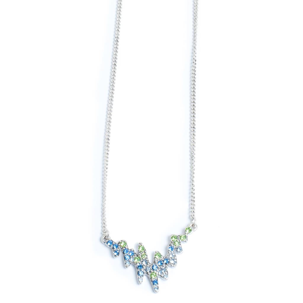 The Northern Lights Necklace by A&C Oslo Norway features a silver chain and a zigzag pendant adorned with blue and green crystals, inspired by Norwegian jewelry.