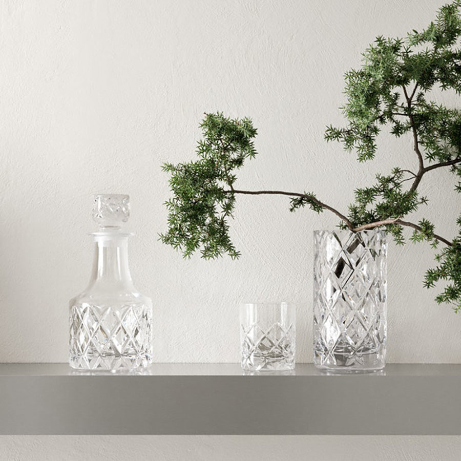 A Scandinavian classic scene: a crystal decanter, glass, and the Orrefors Sofiero Vase Cylinder elegantly displayed on a gray shelf alongside a green leafy branch.