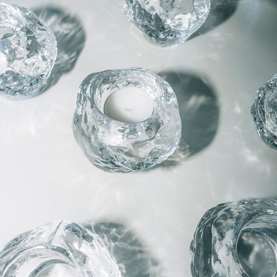 Three clear, irregularly-shaped glass votives from the KostaBoda: Snowball Votive Set of 3 are arranged on a white surface, reflecting light, with a tealight candle glowing in the center holder.