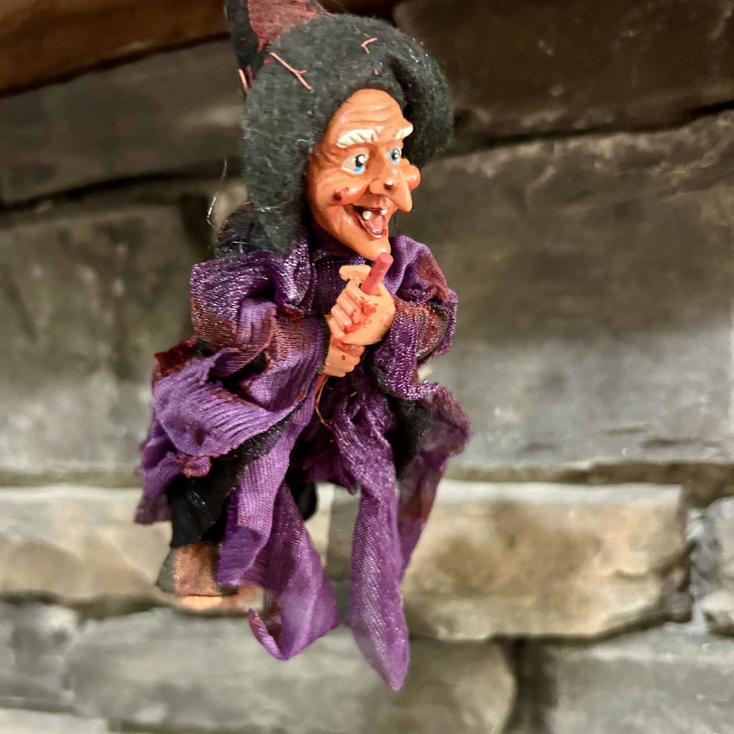 The Walburga Purple Kitchen Witch Doll, handmade in Germany with a pointed hat and purple robe, hangs by a stone wall to bring charm and good luck to your home.
