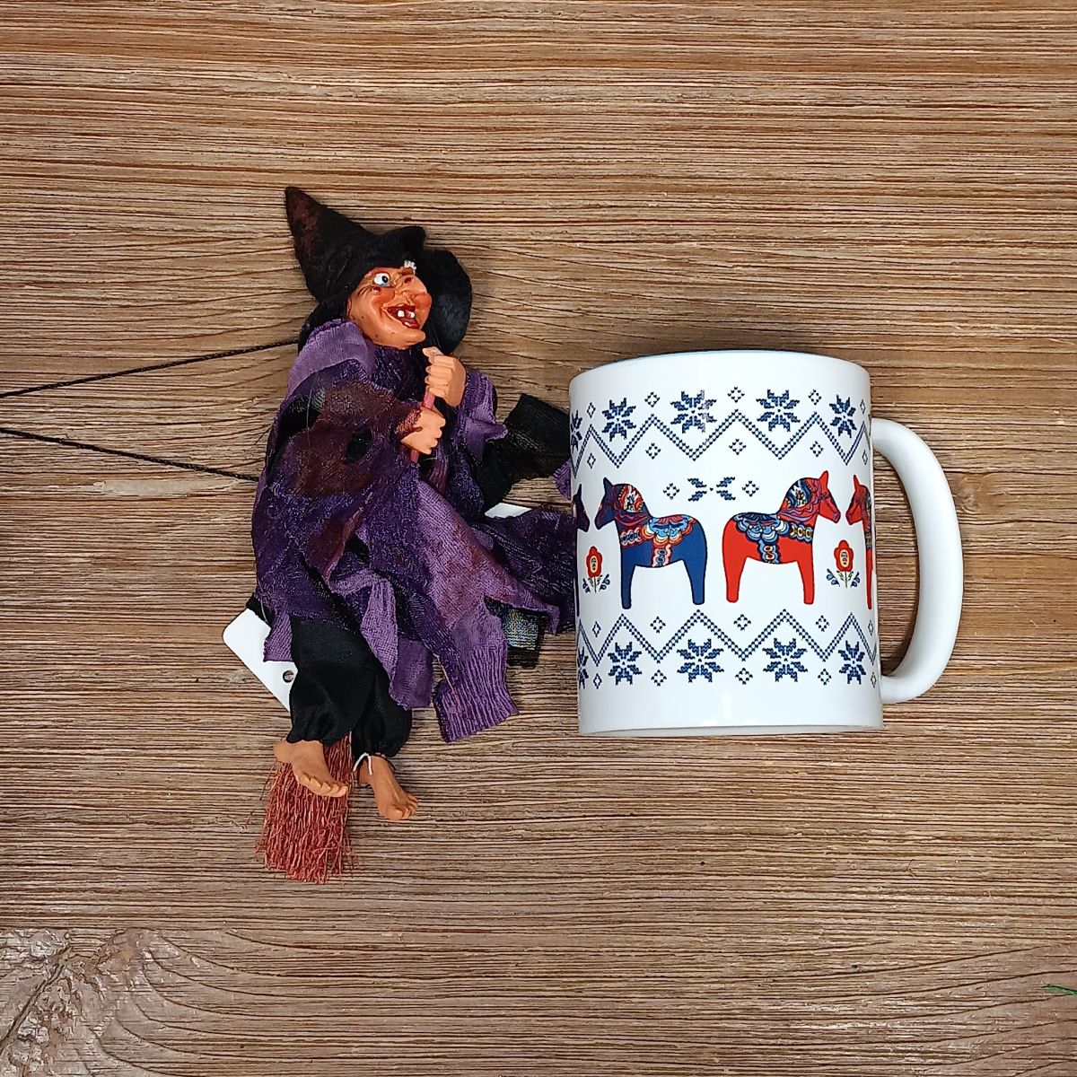 A Walburga Purple Kitchen Witch Doll rests beside a white mug with colorful horse patterns on a wooden surface, serving as a charming good luck charm for kitchen protection.