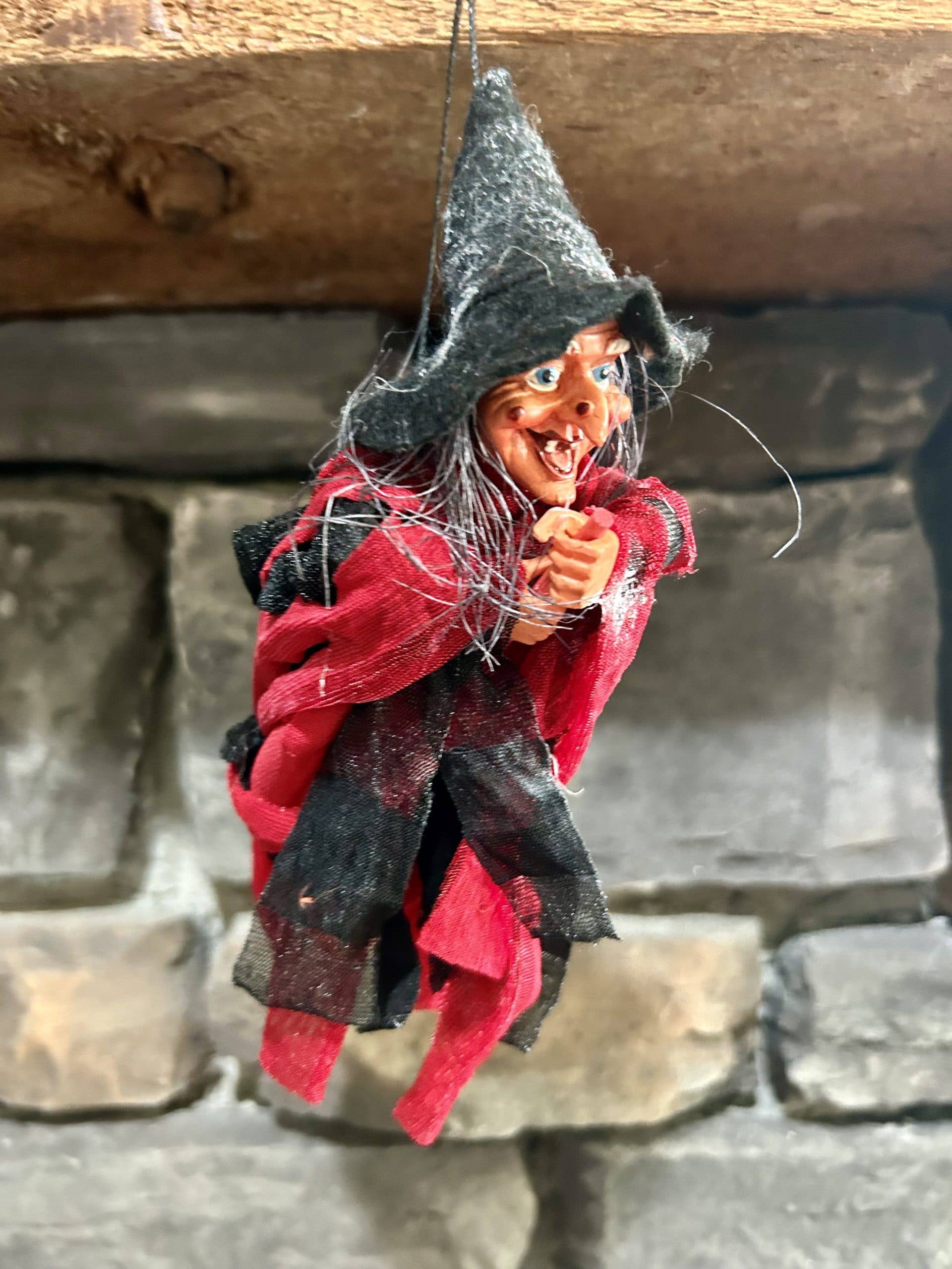 The Walburga Red Kitchen Witch Doll, handmade in Germany and dressed in black and red with a black hat, hangs from a string as a good luck charm against a stone wall background.