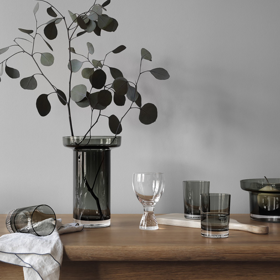A wooden table with dark glass vases, drinking glasses, a KostaBoda Limelight Wine XL Glass 11.8oz (2-Pack) by Goran Warff, a white cloth, and a branch with green leaves sits against a gray wall.