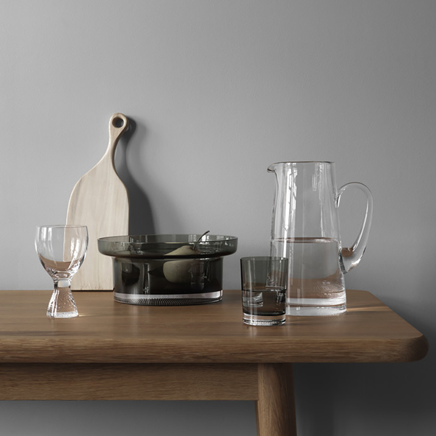 A wooden table holds a glass pitcher, short glass, dark bowl, cutting board, and the KostaBoda: Limelight Wine XL Glass 11.8oz (2-Pack) by Goran Warff from the Kosta Boda collection, all set against a gray wall.