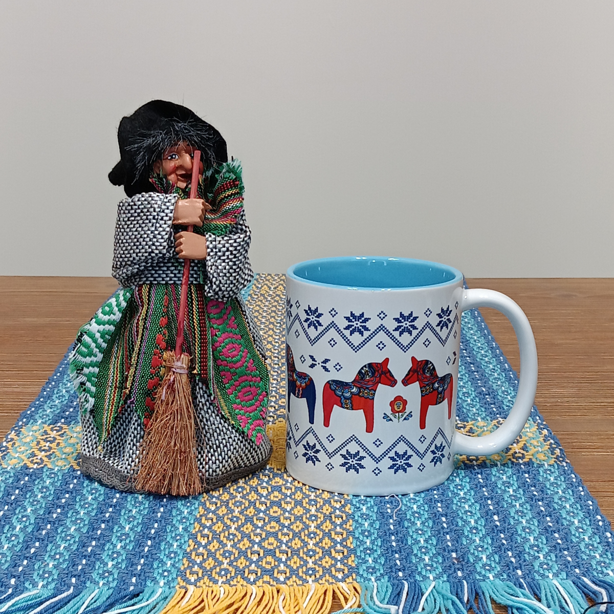 A Yasmina Green Kitchen Witch Doll figurine (7" tall) with a standing base is placed beside a white mug featuring red and blue horses, both set on a woven blue-and-yellow cloth atop a wooden table.