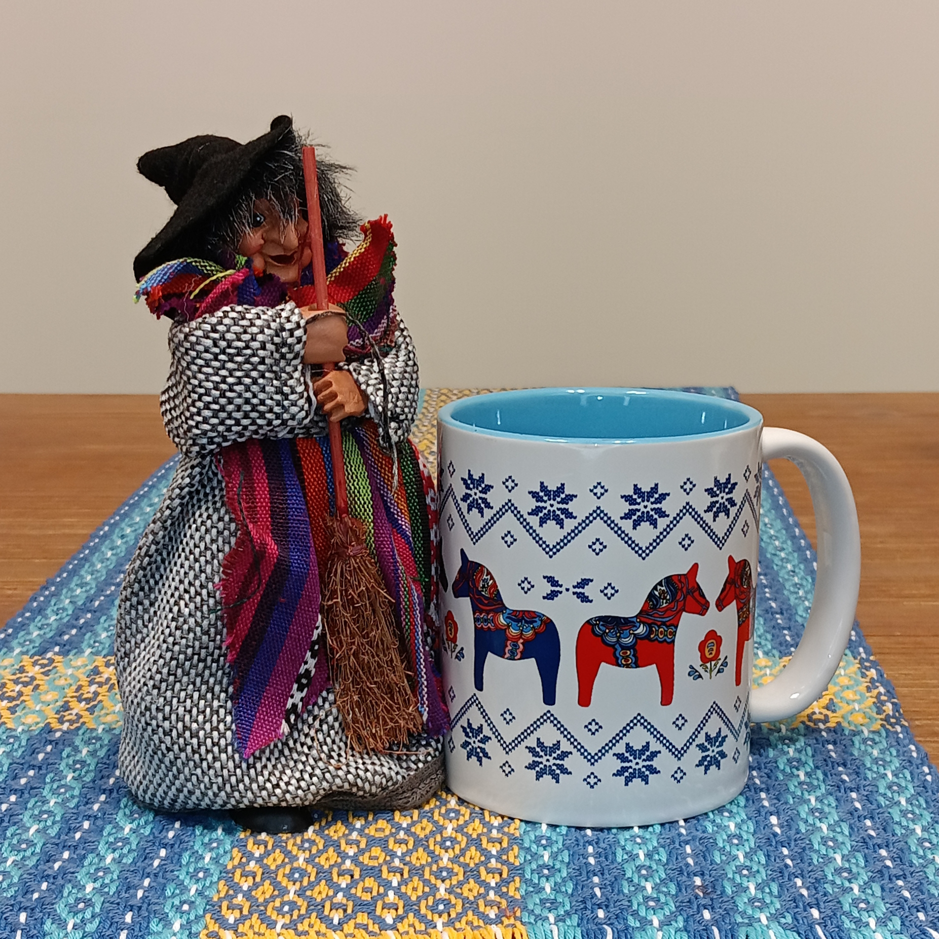 The Kitchen Witch: Yasmina Purple Standing Kitchen Witch Doll with a broom stands beside a white mug featuring colorful Dala horses, set on a blue and yellow woven table runner, bringing Scandinavian folklore charm to your kitchen.