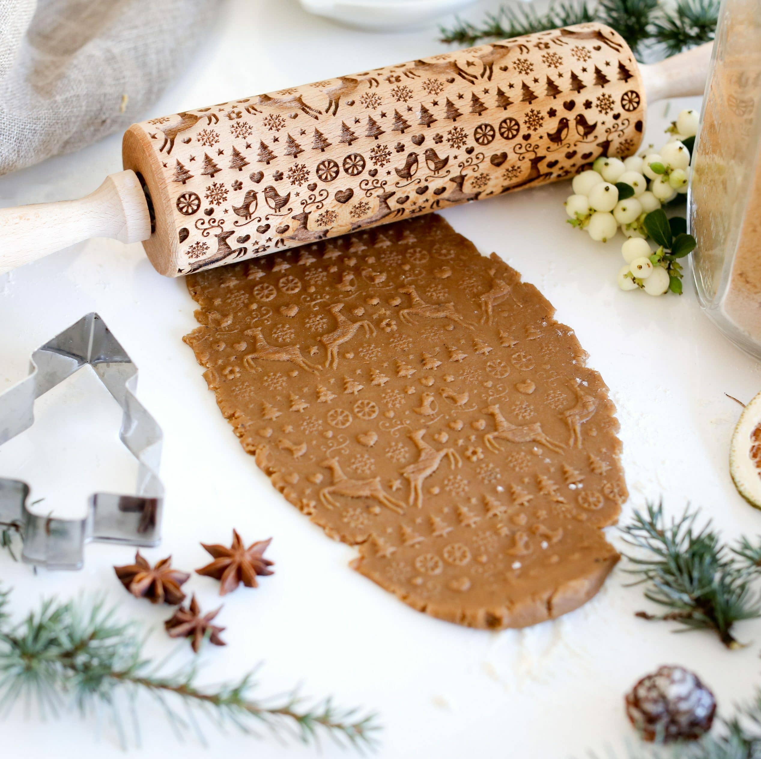 The Rolling Pin: Reindeer Birds & Trees Embossing Rolling Pin, inspired by Scandinavian design and handmade craftsmanship, skillfully embosses gingerbread dough with detailed snowflake and Christmas deer patterns amidst a selection of festive cookie cutters and decorations.
