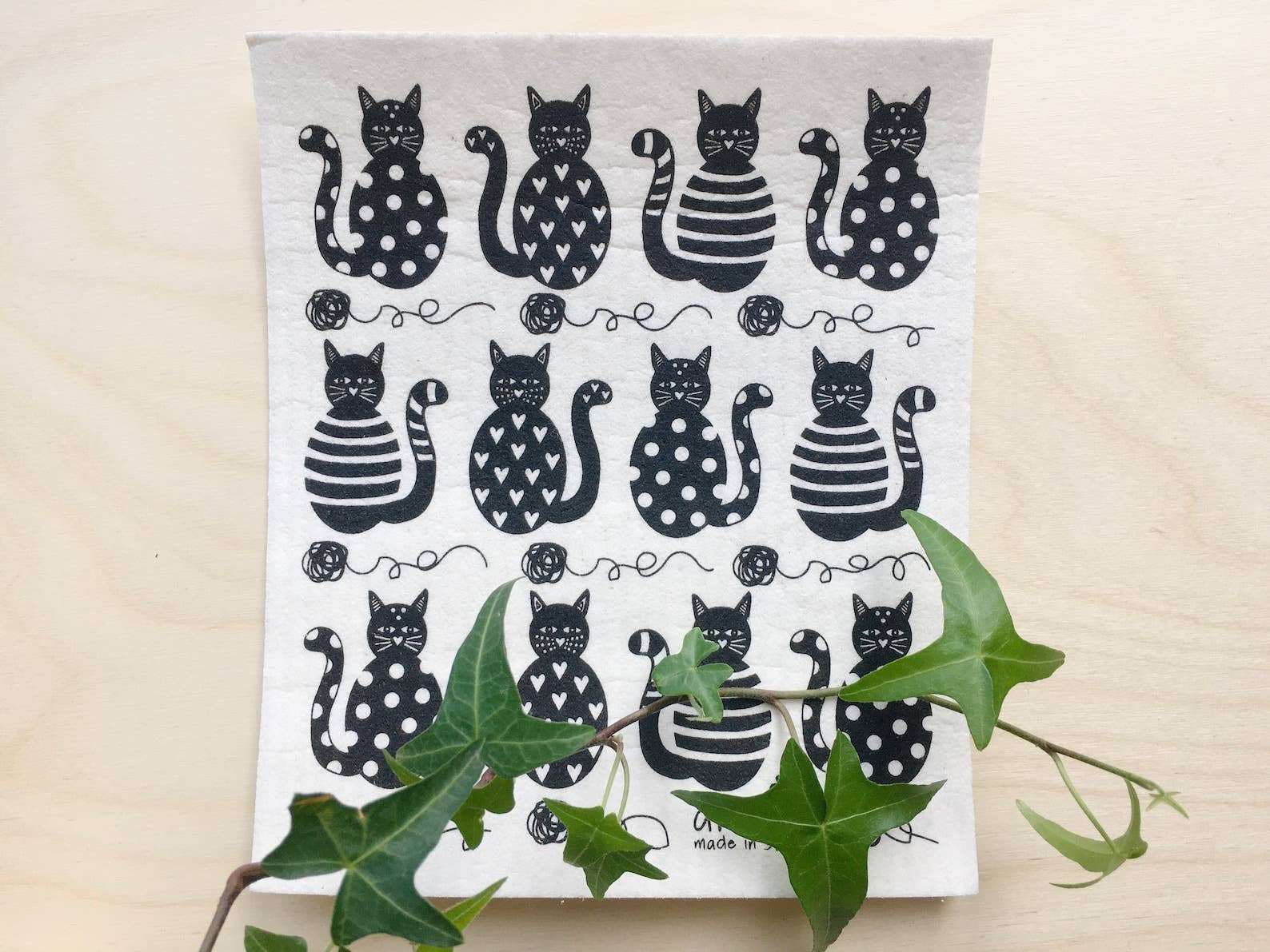 The Dish Cloth: Cats Swedish Dishcloth by Anneko Design Sweden features black cat illustrations in various patterns, with a green ivy plant partly covering the eco-friendly, biodegradable dishcloth's bottom right corner.