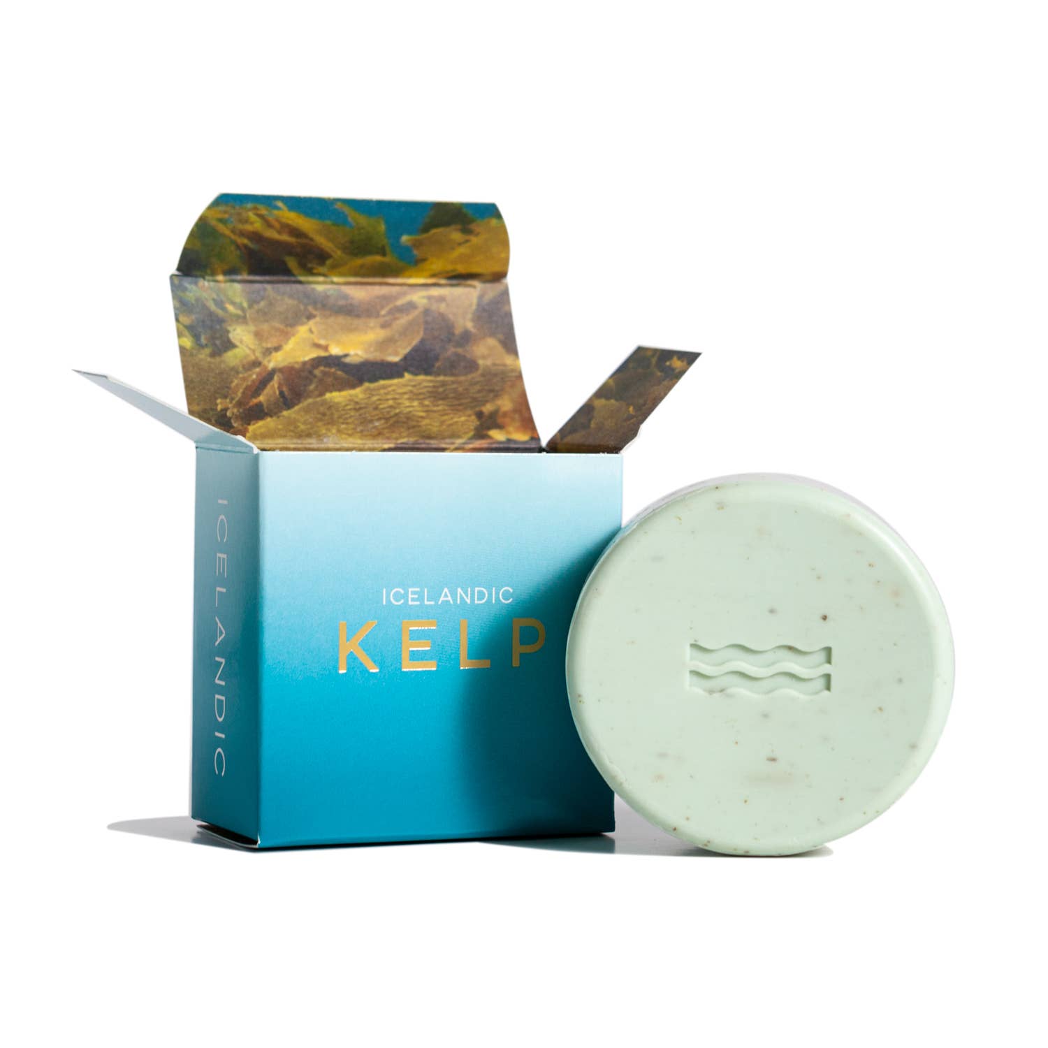A round, green Hallo Iceland Kelp soap bar with a wave design rests beside an open box. Crafted with a sustainable soap base, the packaging features a calming blue and brown color scheme.