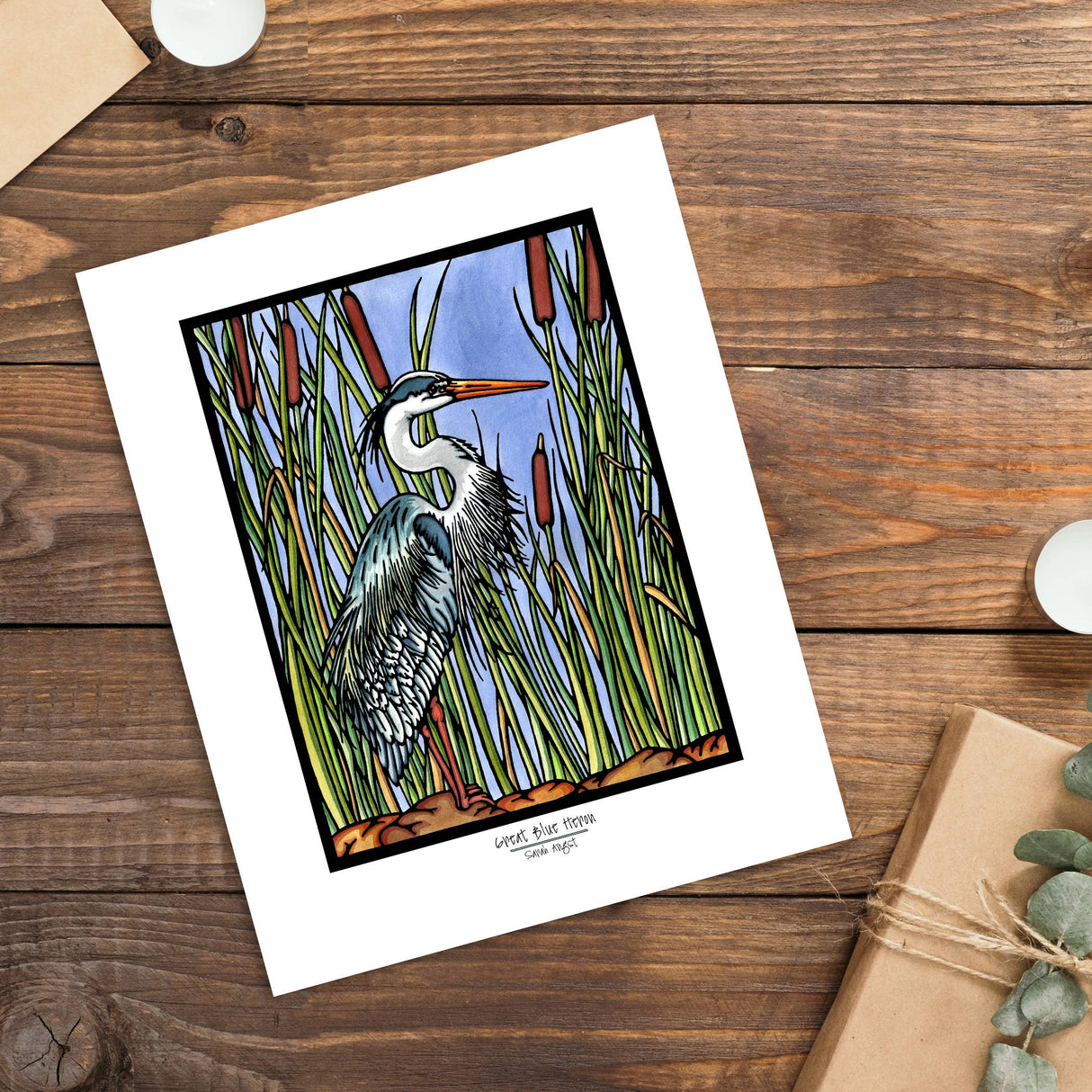 The Artwork: Blue Heron 8x10 Print by Sarah Angst is a giclee print featuring a great blue heron among tall reeds, displayed on wood beside a candle, envelope, and wrapped gift.