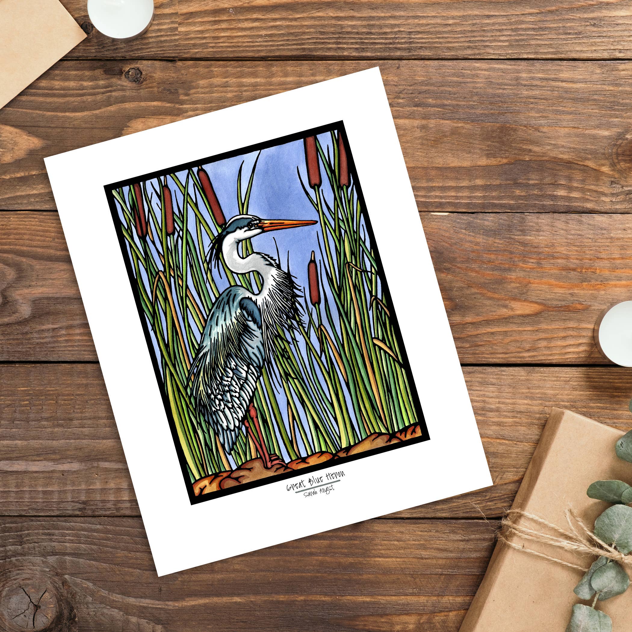 The Artwork: Blue Heron 8x10 Print by Sarah Angst is a giclee print featuring a great blue heron among tall reeds, displayed on wood beside a candle, envelope, and wrapped gift.