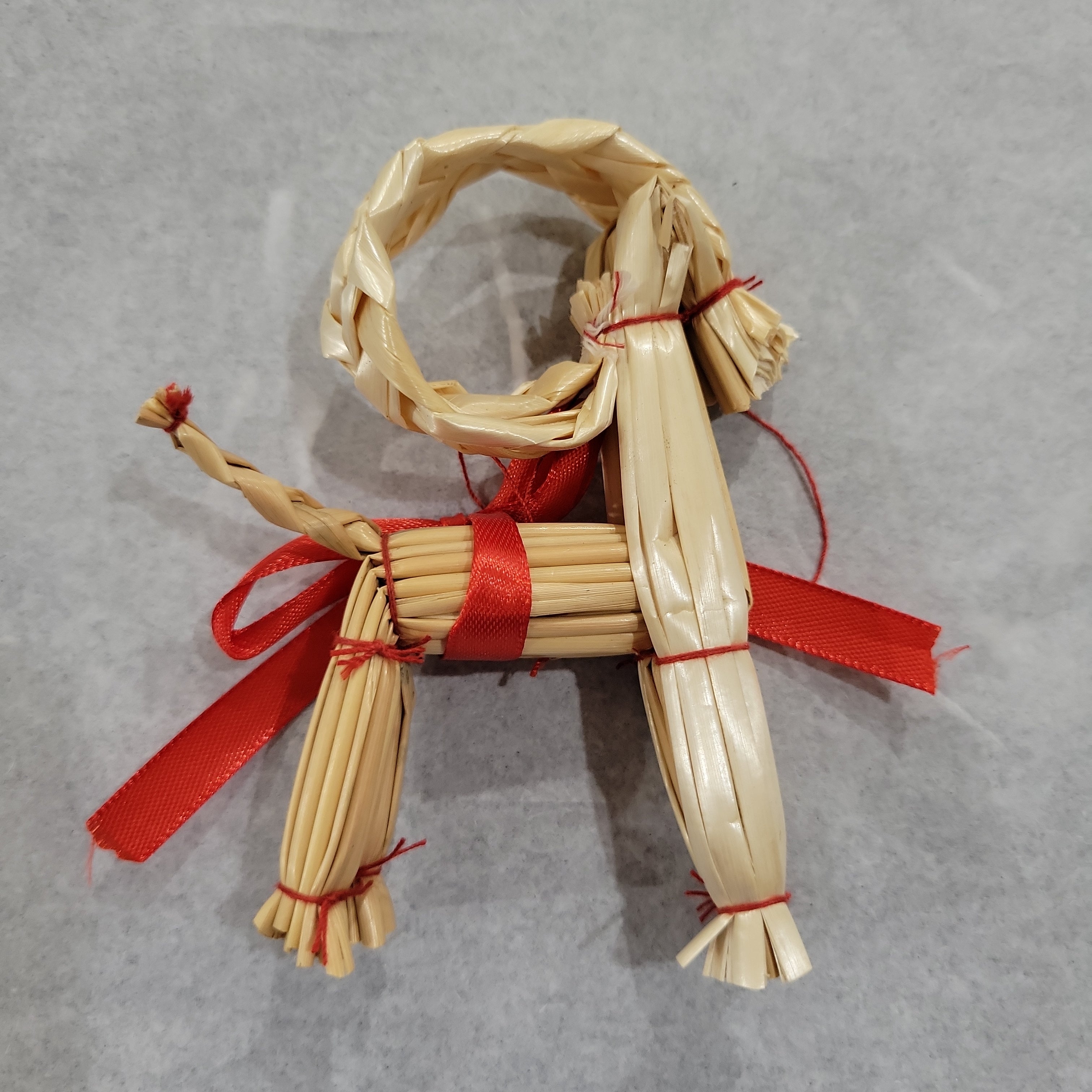 The 4" Straw Goat Julbok Christmas Tree Ornament, handcrafted and tied with red ribbon, sits on a light grey surface—perfect as Nordic holiday décor or unique table decoration.