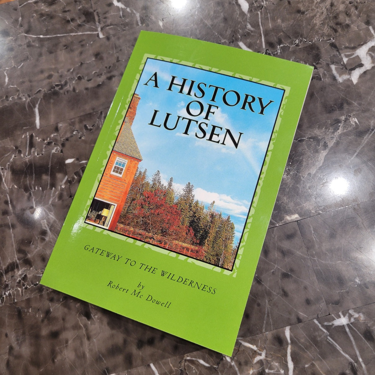An autographed copy of A History of Lutsen by Robert McCain Dowell sits on a marble surface, featuring a green cover with a cabin and trees under a blue sky.