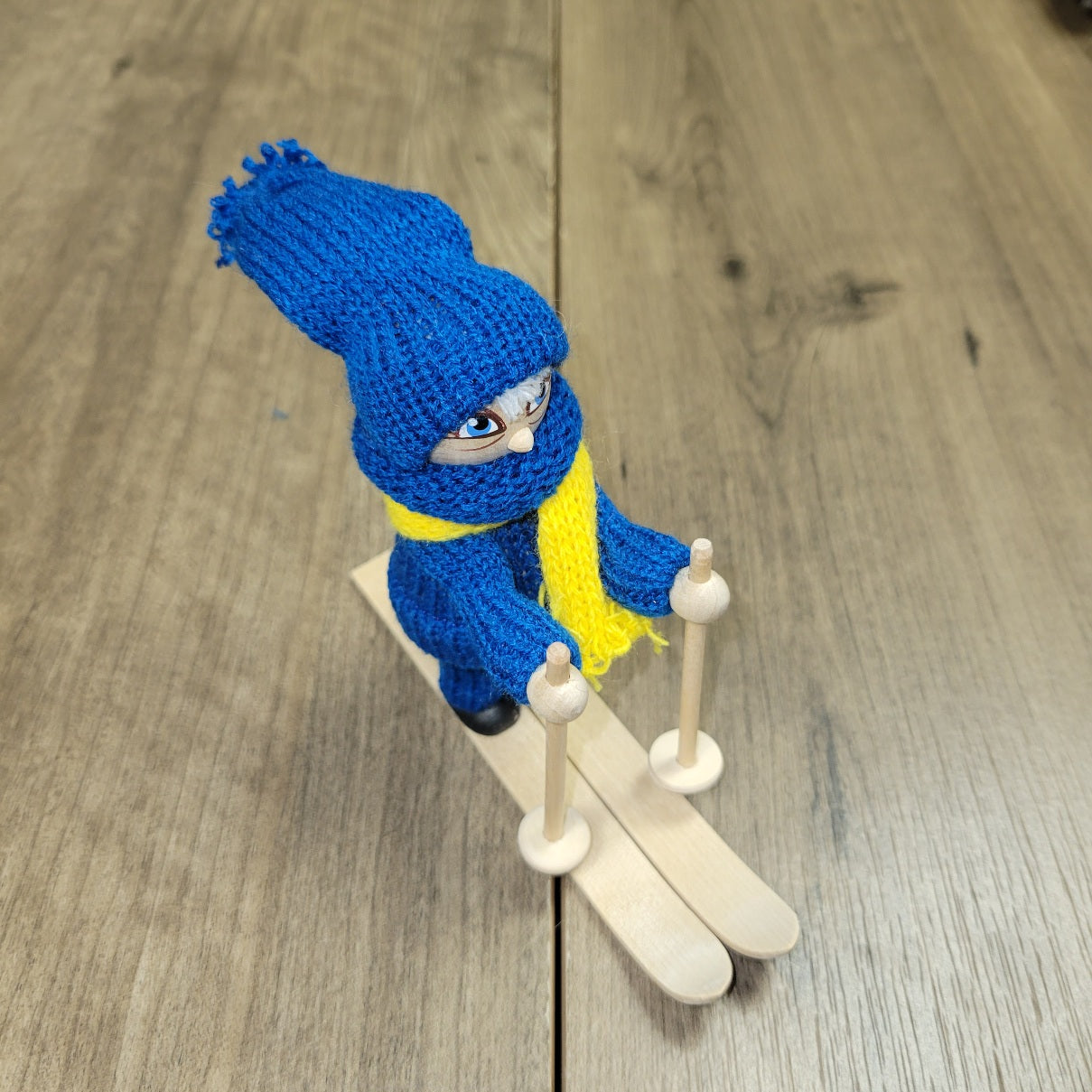The Swedish Skier Figurine, hand crafted in Sweden, features blue and yellow attire, stands 4.25" tall on 5" long skis, and holds ski poles on a wooden base.