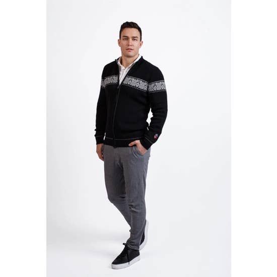 A man stands against a white background, wearing the Breheimen Merino Zip Cardigan in black with a Nordic-inspired pattern, gray pants, and black sneakers. One hand is in his pocket.