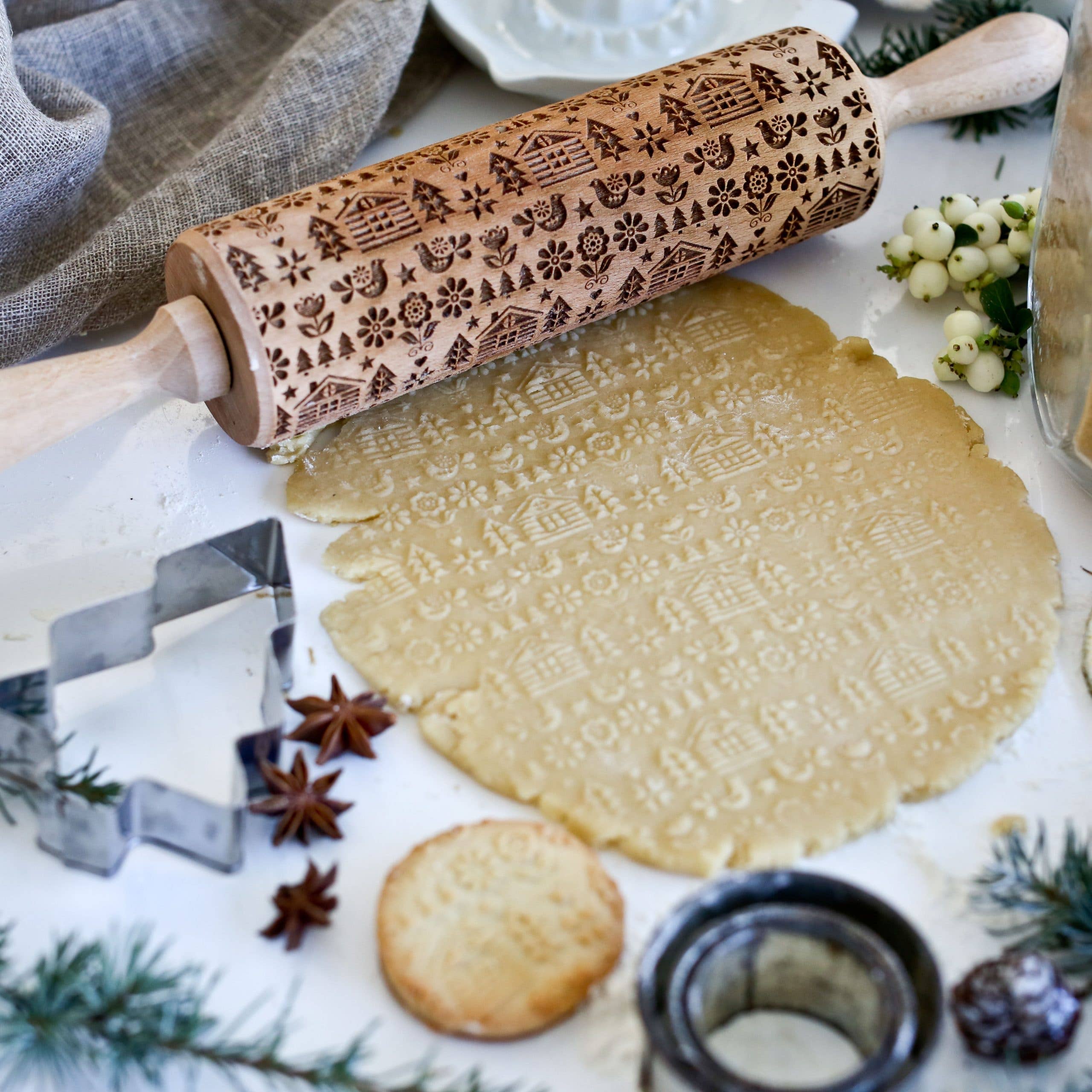 Festive decorations, baking tools, and spices surround cookie dough imprinted with the Folk Pattern Winter Cabin embossing rolling pin, creating a delightful holiday scene on a white surface. This Scandinavian-style pin brings an artisanal touch to the charming tableau.